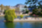 People relax along the riverbank under a willow tree, with Parisian buildings in the background. Clear blue sky and green foliage set a calm mood.