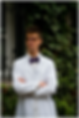 Will Inrig in white lab coat and bow tie stands with arms crossed in front of ivy-covered wall, looking thoughtful and composed.