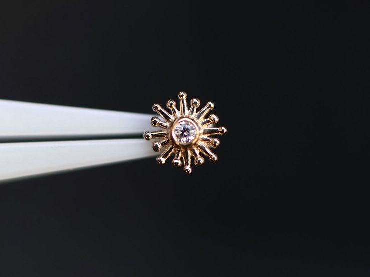 A solid 14k gold sunburst top with a clear centre stone, held in tweezers against a dark background.
