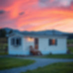 White mobile home with a wooden door and lit porch light, set in a lush field. Vibrant pink and orange sunset sky, mountains in background.