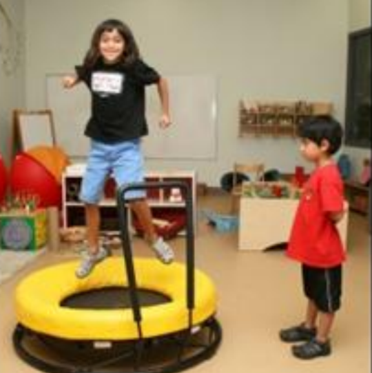 Child happily jumping on yellow trampoline indoors, another child watches closely.