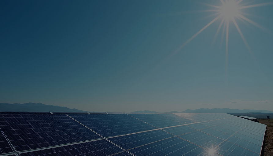 The image shows a vast field of solar panels stretching across the foreground under a clear, sun-drenched sky with hazy mountains visible in the distant background.