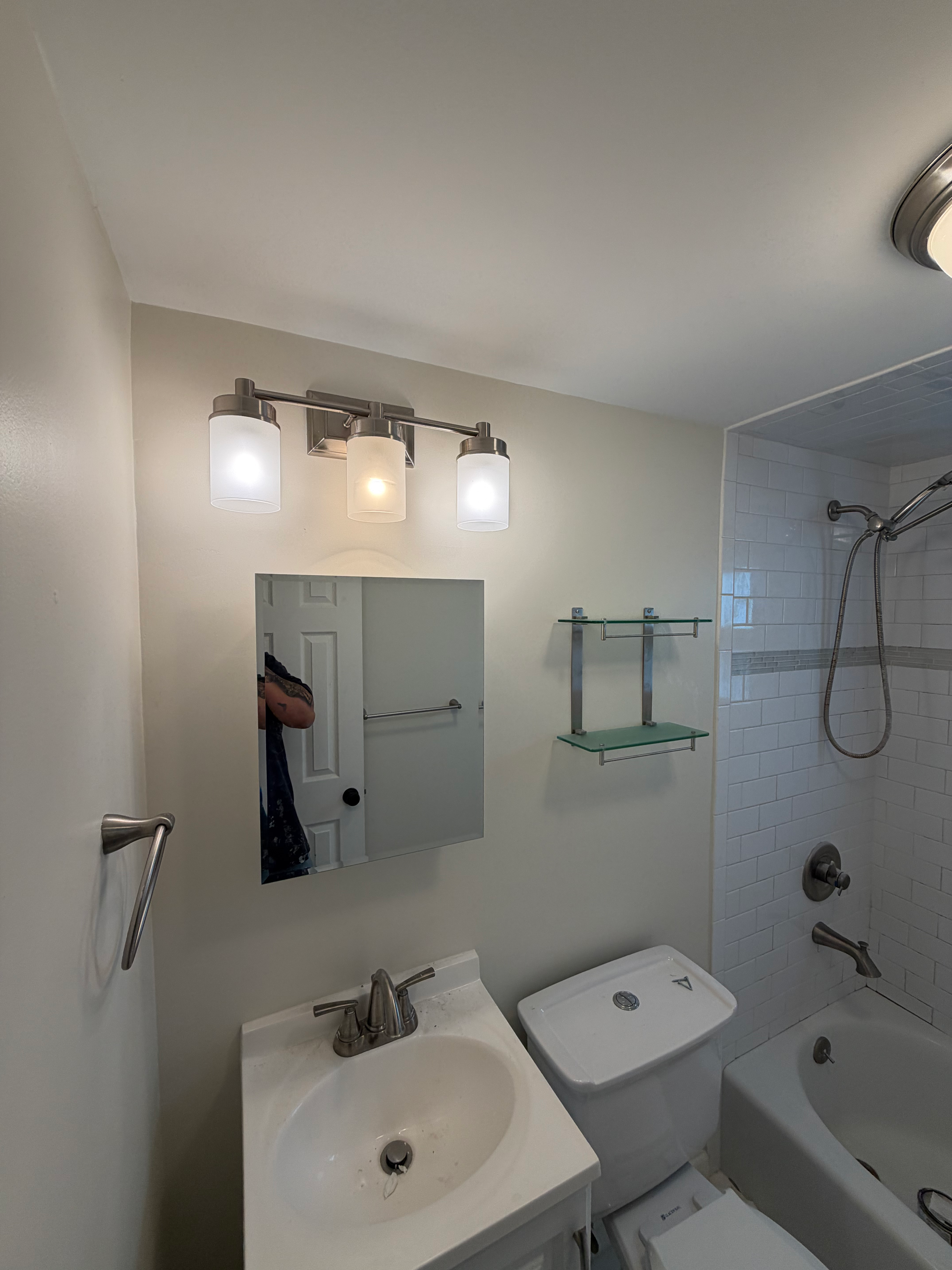 Small bathroom remodel finished in Bergen County, NJ — white subway tile tub surround, vanity with mirror cabinet, and glass shelving