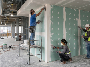 Workers in construction gear apply joint compound to drywall in a large, unfinished room. Ladders, buckets, and tools are scattered around.