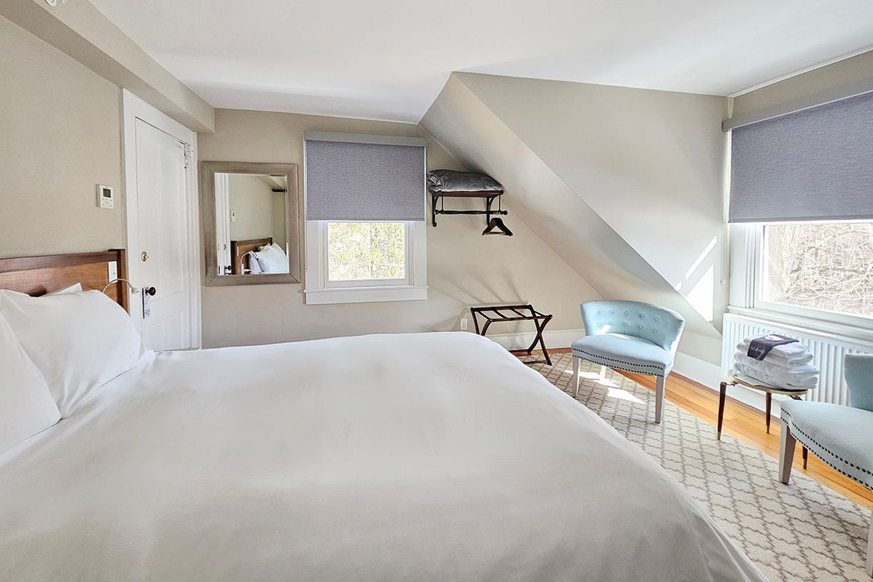 Room 32 at Lakehouse Inn featuring a cozy queen bed, shared balcony with sunrise and lake views, classic inn charm with natural light, and additional seating for a relaxing stay