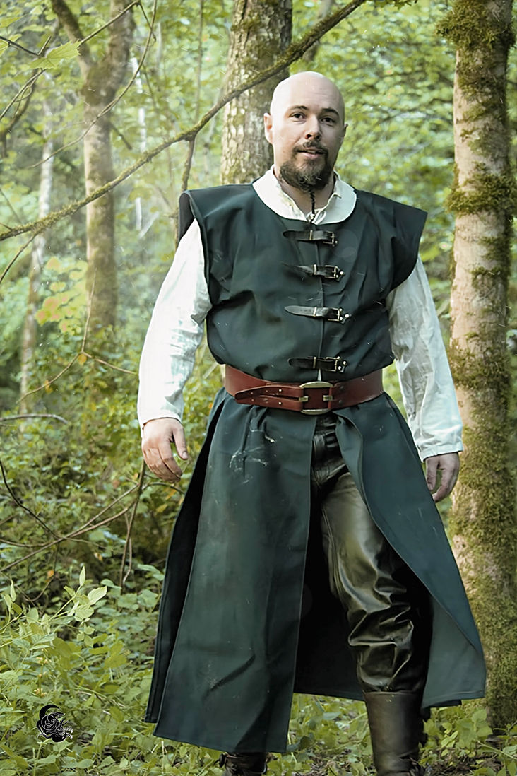 Homme barbu porte une tunique verte et regarde la caméra dans la forêt.