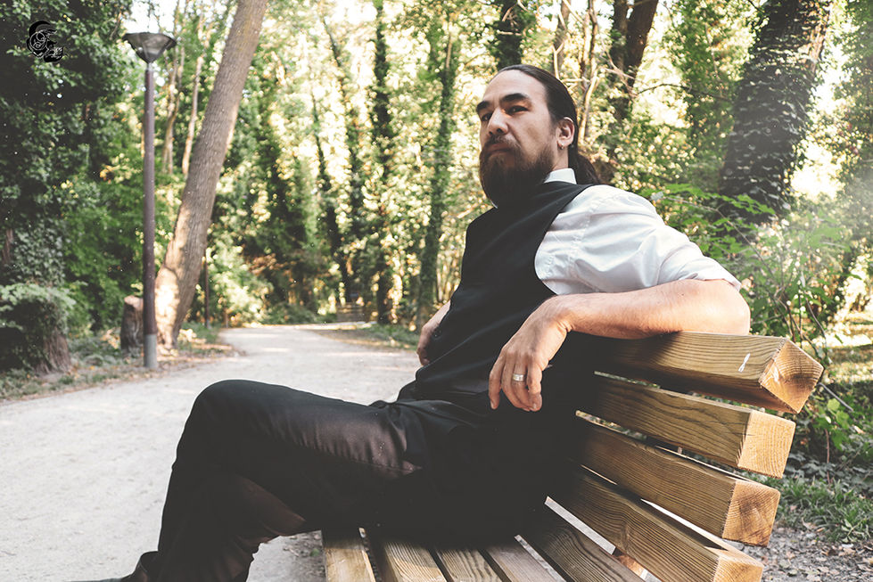 Un homme barbu assis sur un banc, regardant loin, en arrière-plan, en plein air.