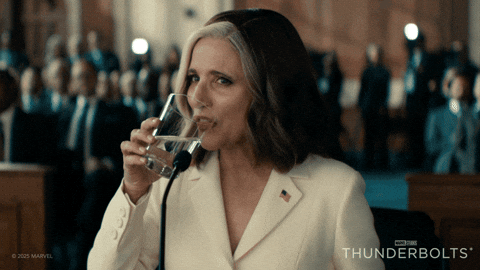 A woman in a white suit drinks water confidently in a courtroom setting. A crowd is blurred in the background. Text: "THUNDERBOLTS."