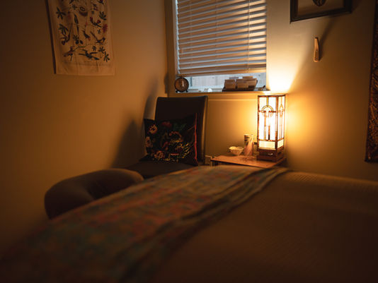 A massage table and chair in warm yellow light. A window is visible in the background with the shades mostly drawn.