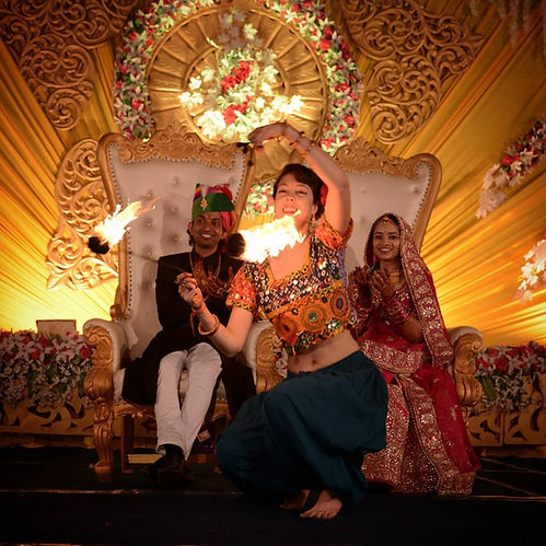 Fire dancer mystica fiora spinning fire poi on stage with the bride and groom at an Indian wedding reception party event surrounded by decorations and ornate designs in a sparkly cultural costume belly dance costume and audience is smiling happy exhilerated