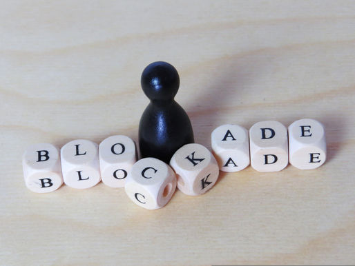 A black game-piece figure stands among lettered cube beads spelling “BLOCKADE,” with scattered and tilted cubes creating the impression of an obstacle or disruption.