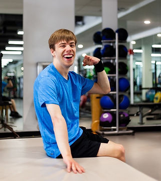 Young man at the gym. He is wearing a blue workout shirt and black shorts and is smiling, sitting, and flexing his bicep.