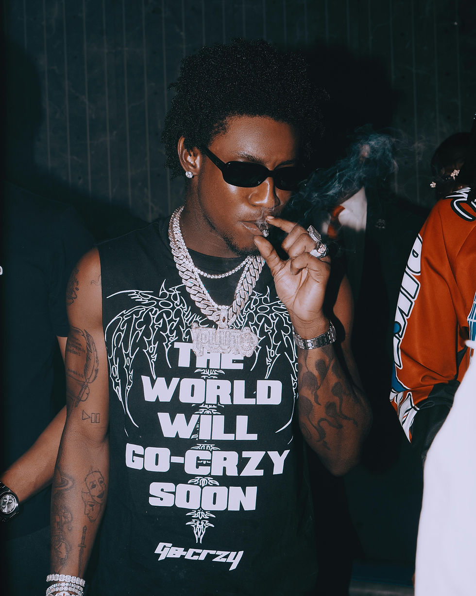 Person in sunglasses and chains smokes in a dark setting. Black shirt reads "THE WORLD WILL GO-CRZY SOON." Relaxed mood with visible tattoos.
