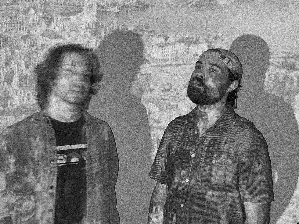Two men stand with projections on them, one looks blurry; background shows a cityscape. Black and white, moody atmosphere.