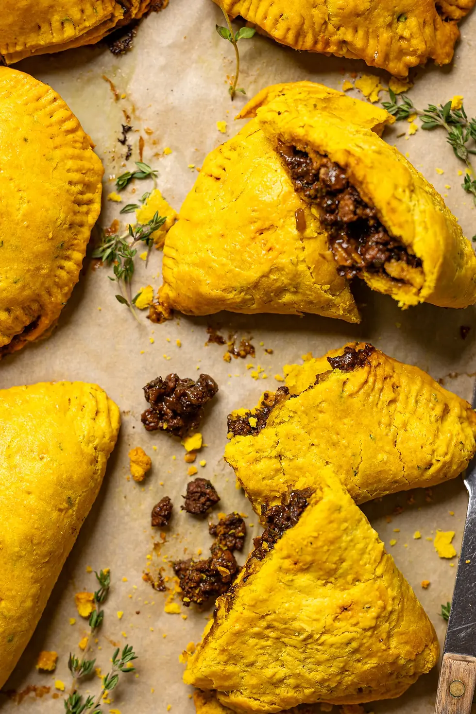 Golden pastries filled with spicy meat, crumbled on parchment. Vibrant yellow crust, scattered thyme sprigs, rustic and appetizing.
