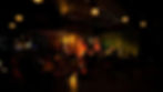 Musicians perform on stage with guitars and bass in a dimly lit venue. Audience seated on orange chairs. Warm, cozy atmosphere.