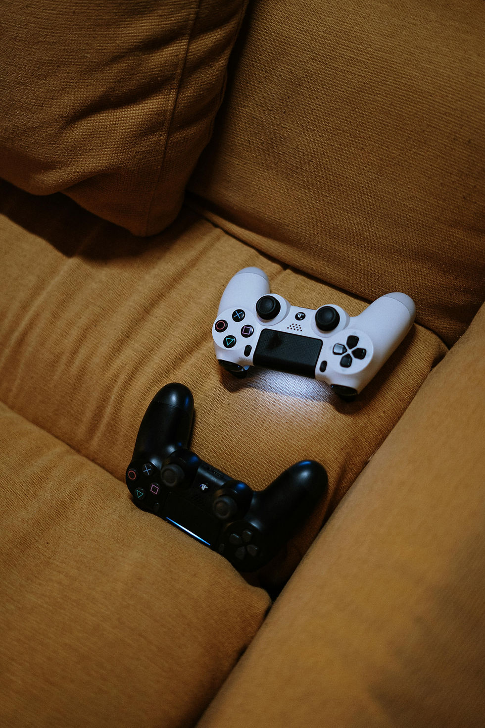 Two gaming controllers, one white and one black, sit on a light brown sofa. The setting is cozy, with a relaxed atmosphere.