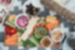 A wooden board with colorful veggies, crackers, dips, and fruits on a patterned cloth. Bowls of olives and chutney are nearby. Casual picnic vibe.