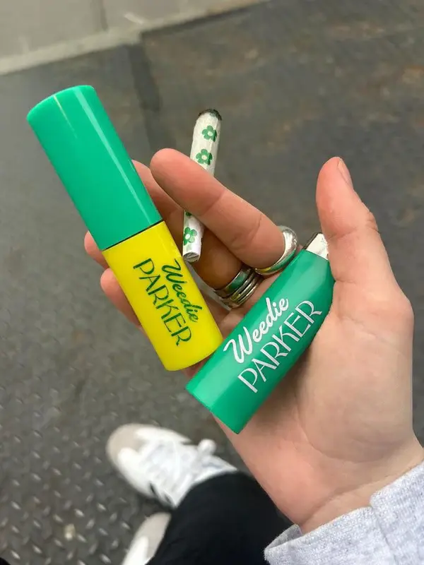 Hand holding a green lighter and yellow lipstick with "Weedie Parker" text, plus a cigarette with green flower pattern, outdoors.