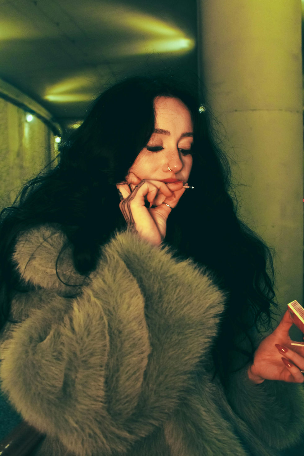 Woman with long hair and nose ring lights cigarette. She's wearing a fur coat in a dimly lit tunnel, holding a matchbox, with a relaxed expression.