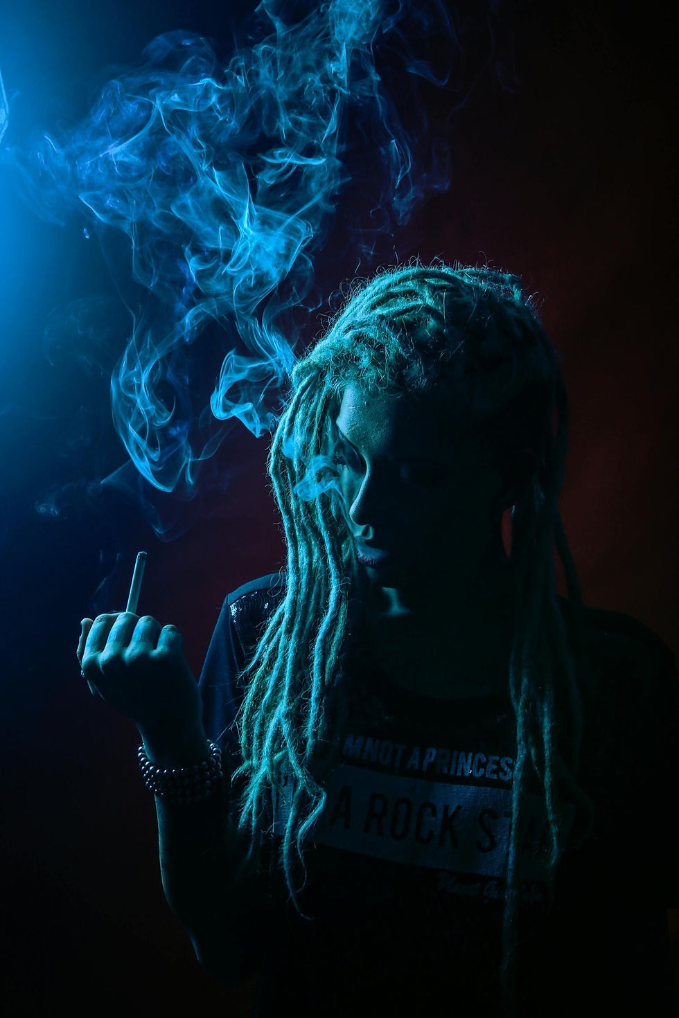 Person with dreadlocks holds a cigarette, surrounded by blue smoke. Dark background, text on shirt reads "I'm Not a Princess, I'm a Rock Star."