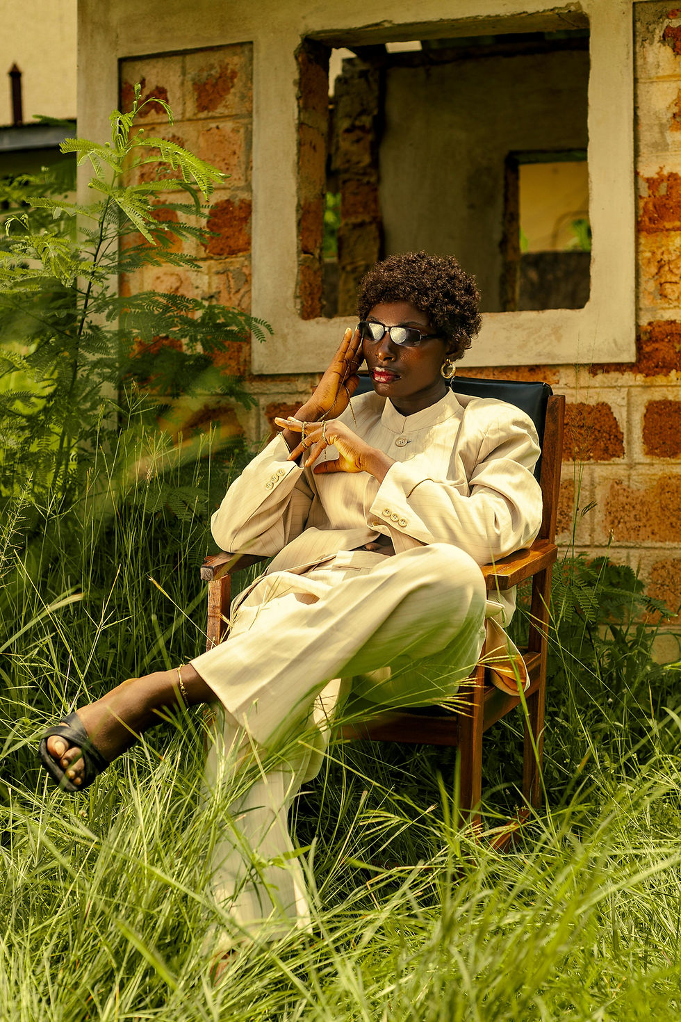 Person in a beige suit sits in a chair surrounded by tall grass, leaning pensively on their hand. Brick building with window in background.