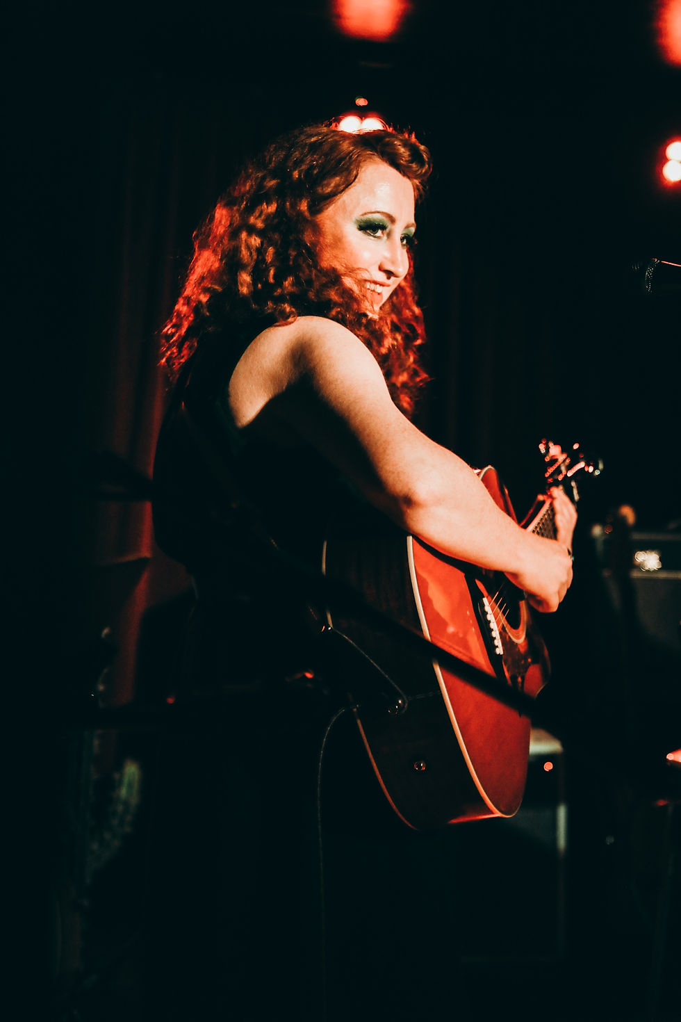 A person with curly red hair smiles while playing an acoustic guitar on stage. Warm lighting creates a cozy, energetic atmosphere.