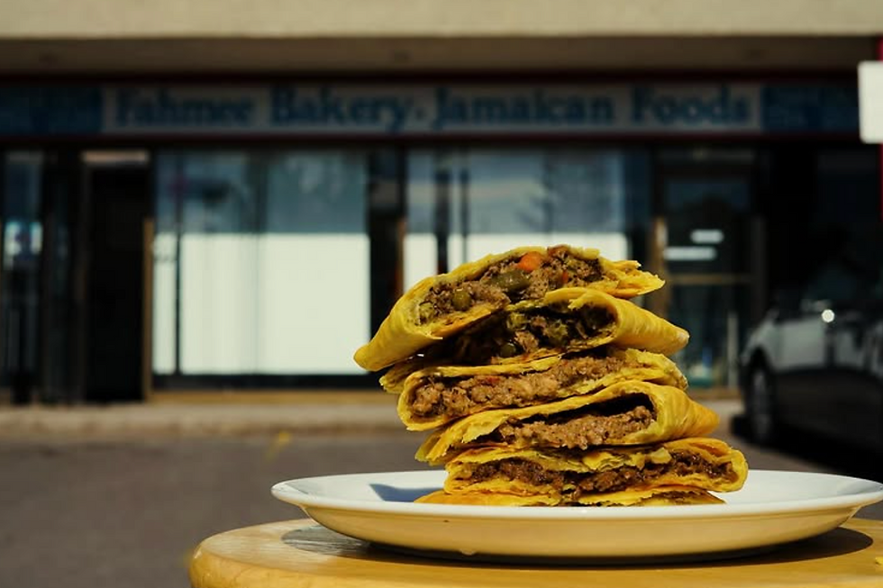 Stack of meat-filled pastries on a plate in front of a bakery. The sign reads "Fahmee Bakery, Jamaican Foods." Warm, sunny setting.