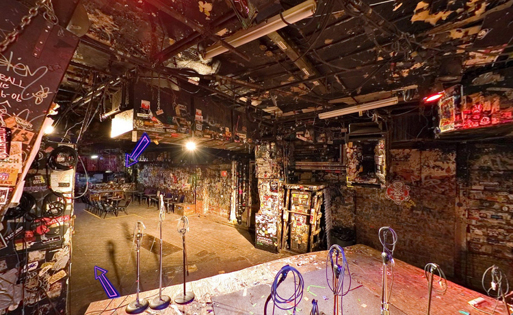 Underground music venue with graffiti-covered walls, empty microphone stands on stage, and dim lighting. Tables and chairs are scattered around.