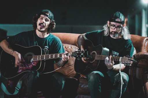 Two men play guitars on a brown couch, one singing. Both wear caps and the mood is relaxed. Dimly lit indoor setting.