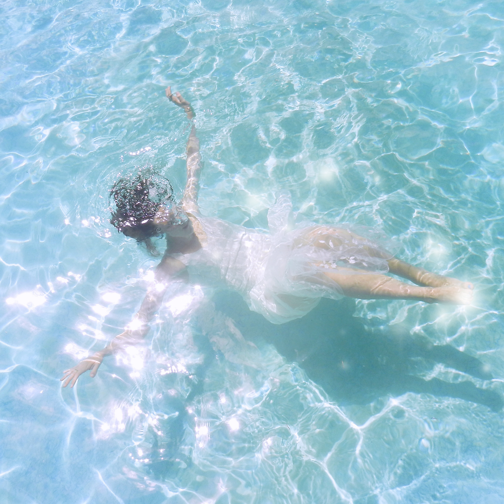 Person floating in clear blue water, wearing light clothing. Sunlight sparkles on the water, creating a tranquil, dreamy atmosphere. No text.