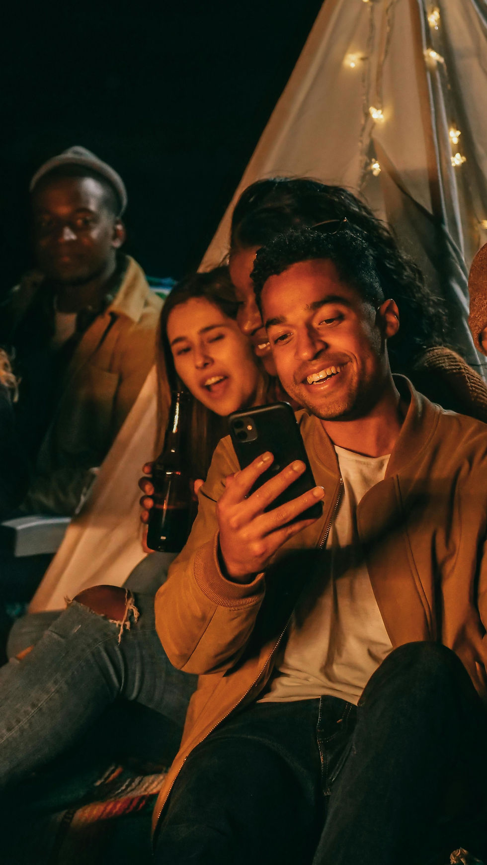 Group of friends at night, sitting outside a tent with lights, smiling at a smartphone. One holds a beer bottle. Cozy and joyful mood.