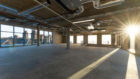 Open-plan interior with exposed ceiling services, concrete floor, and large windows in natural light