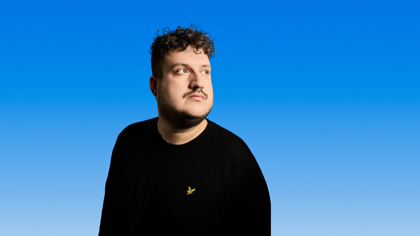 Portrait von Robin ab der Brust aufwärts, der Hintergrund ist ein blauer Verlauf. Er guckt mit neutralem Blick rechts aus dem Bild.