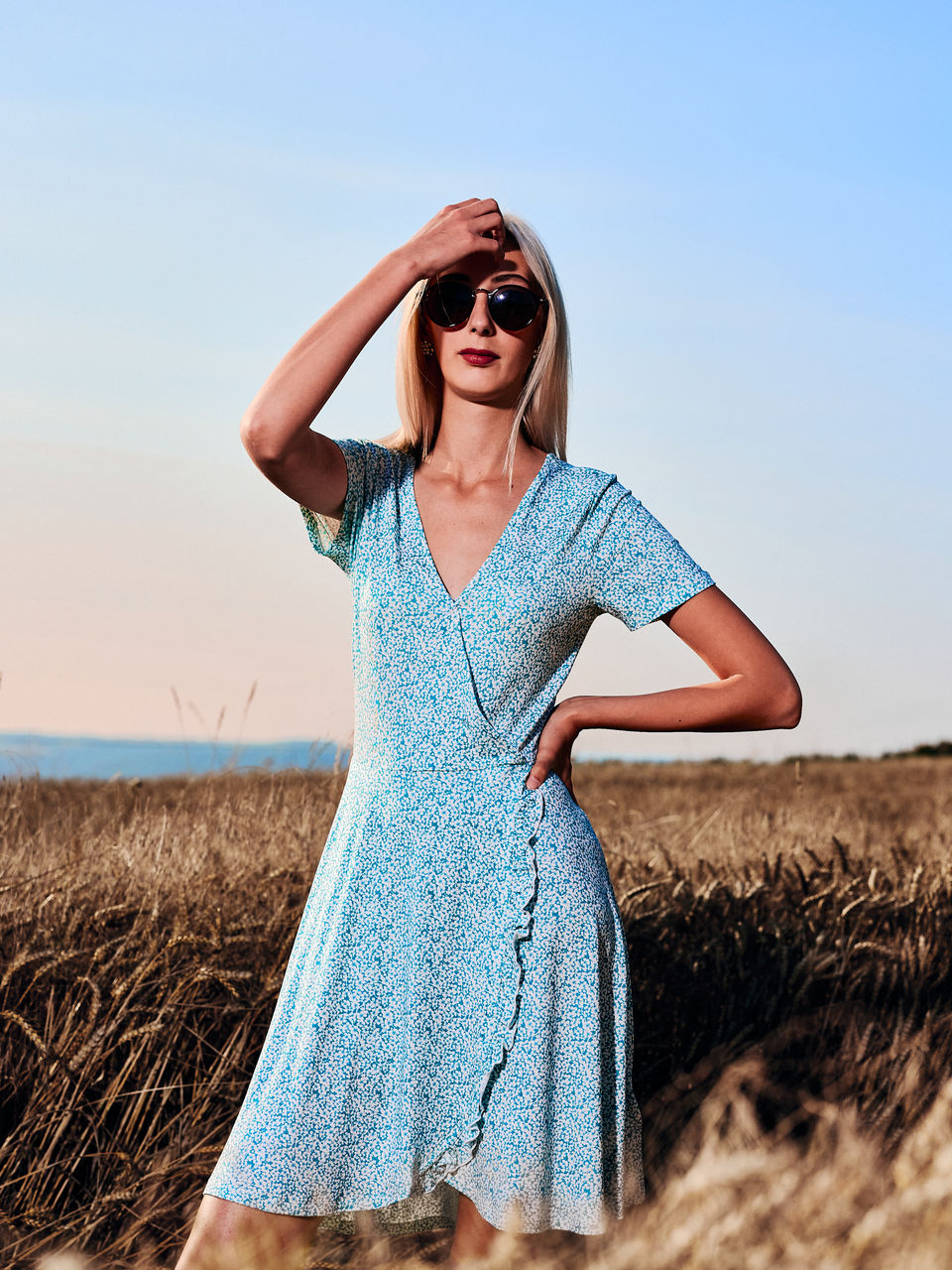 Ein Fashion-Portrait von Christin in einem Feld im Spätsommer. Sie trägt ein blaues Kleid sowie eine Sonnenbrille. Die Ähren sind golden und liefern einen schönen Kontrast zum Blau. Sie ist mit Blitzlicht beleuchtet und steht so klar im Fokus.