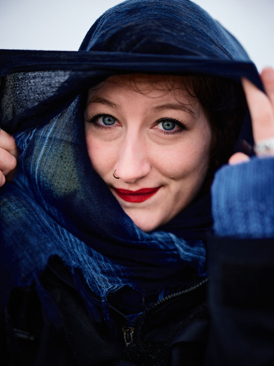 Ein nah aufgenommenes Portrait von Pia im Regen. Sie schützt sich mit einem Blauen Schal der ihr Gesicht einrahmt. Sie blickt den Betrachter direkt an, ihre blauen Augen und roten Lippen sind der Eyecatcher. Sie lächelt leicht.