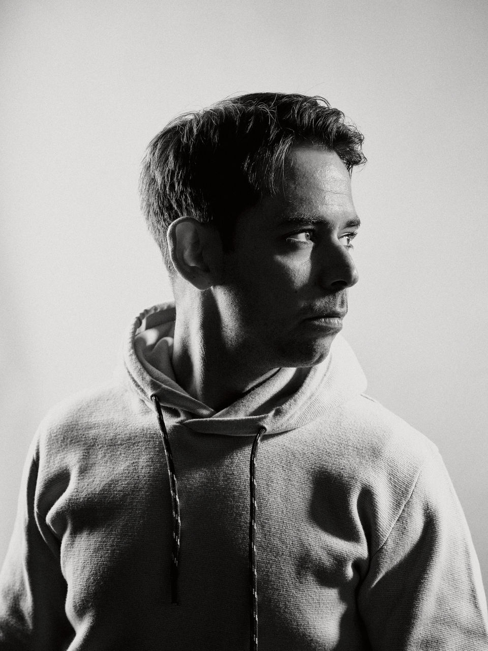 Portrait eines Mannes, der Ausschnitt zeigt eine Büste von der Brust bis über den Kopf. Er steht frontal zur Kamera, wobei er über seine Schulter nach rechts aus dem Bild heraus guckt. Das Foto ist in Schwarz-Weiß gehalten und hat einen starken Kontrast. 