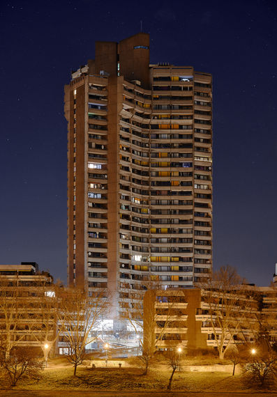 Ein bewohntes Hochhaus in Mannheim im Stile des Brutalismus. Die Straßenlaternen werfen oranges Licht am Boden während der Himmel tief blau ist.