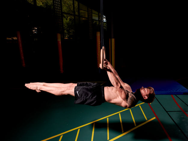 Ein Turner hängt im Plank an den Ringen. Man kann die Muskulatur und Anspannung deutlich sehen. Der Hintergrund ist im Low-Key-Stil, man sieht nur den Hallenboden. 