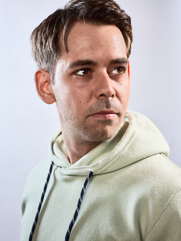 Headshot Portrait eines Mannes. Er ist von der Seite gezeigt wobei er über seine Schulter nach rechts aus dem Bild heraus guckt. Das Foto ist in Farbe und mehreren Blitzen beleuchtet, der Look ist natürlich gehalten.