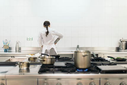 Female private chef standing in a large kitchen cooking on a stove . In the front of the photo is another stove with large stock pot and small pot. Dishes and cooking utensils are placed on the counter near the stove