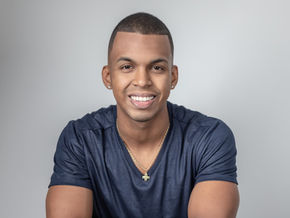 Man with short hair smiling warmly in a studio headshot while wearing a navy shirt and necklace against a light background.