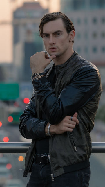 Man wearing a black jacket standing on an overpass at dusk with blurred city lights and buildings in the background