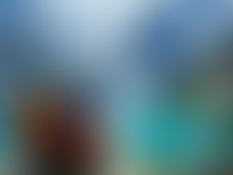 Two hikers with backpacks sit on a rock overlooking a stunning turquoise alpine lake surrounded by tall pine trees and snow-capped mountains under a partly cloudy sky.