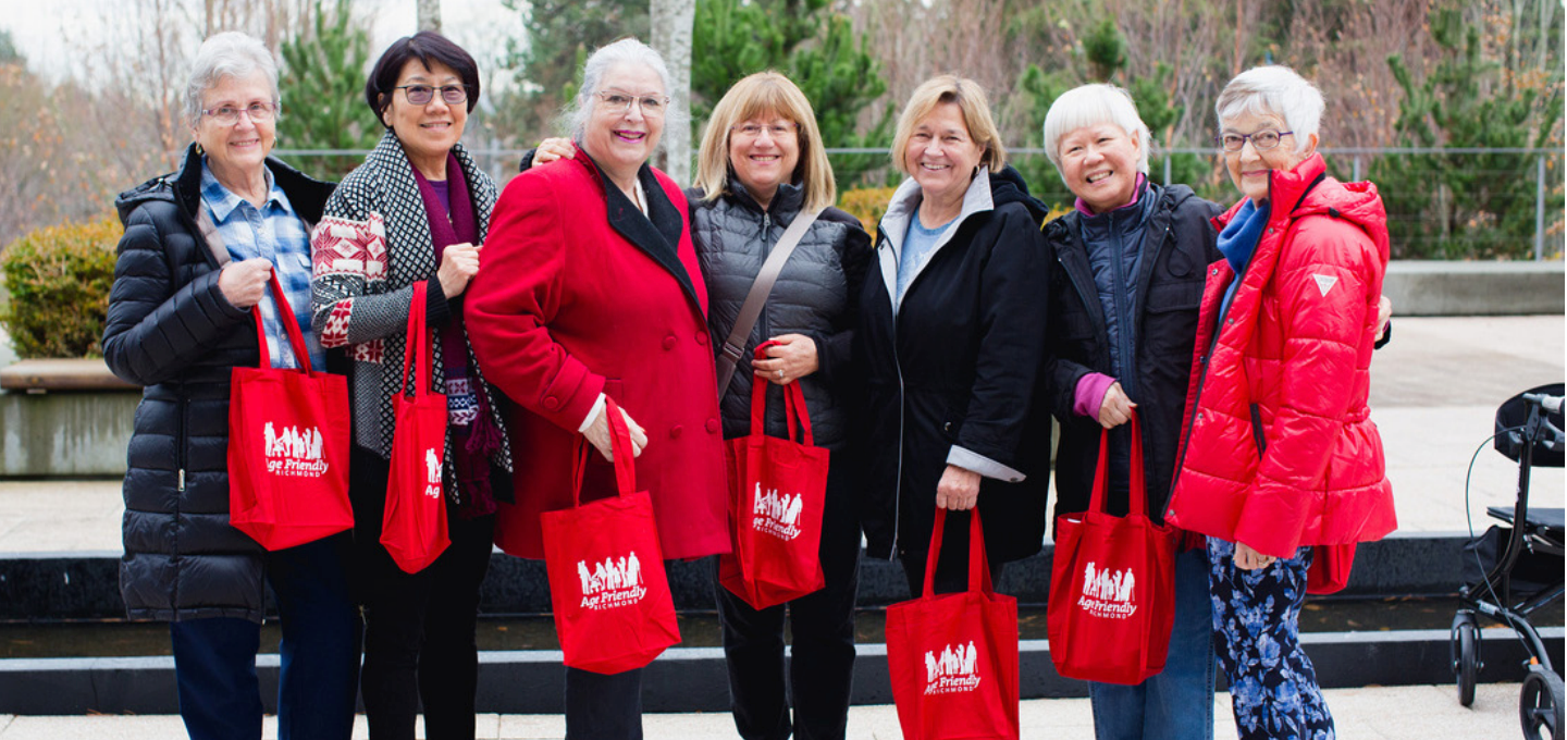 A group photo of a seven senior women carrying a red tote bags from the Age Friendly program.