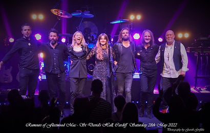 A band of seven musicians stand together to take a bow onstage, standing next to each other in a line, with brightly coloured stage lights behind them.