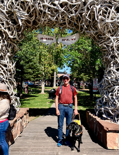 Guest and their guid dog pose on a sunny day under the Antler Arch at Jackson Hole