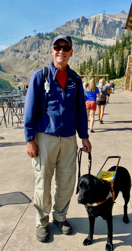 Guest poses in front of an impressive mountain view with their guide dog on a sunny day