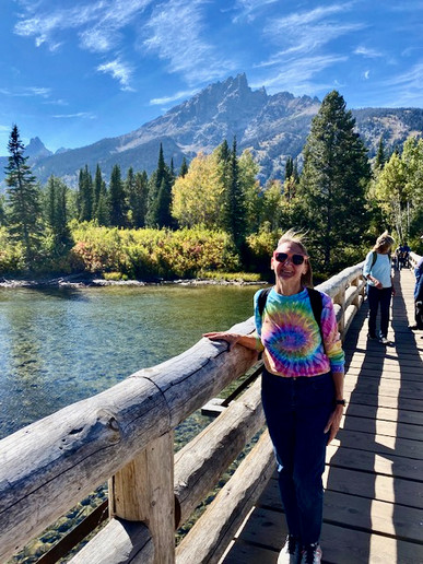 A guest poses in the sun on a footbridge traversing a bright lake in front of the mountains in Jackson Hole