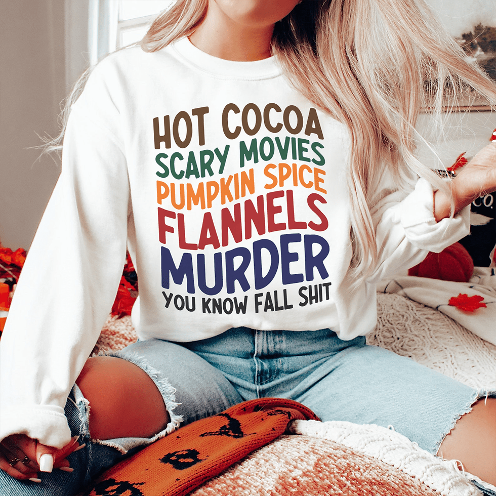 Woman wearing a white sweatshirt with "Hot Cocoa, Scary Movies, Flannels, Murder" on it.